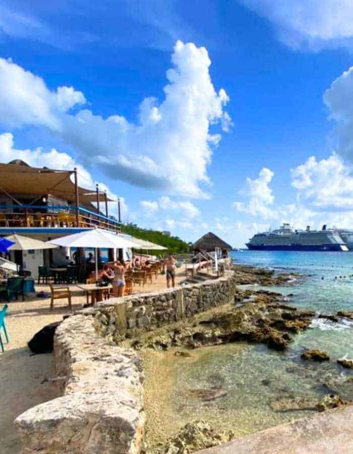 Sunset Beach in Cozumel with a stone seawall and open-air restaurant overlooking clear turquoise water and a rocky shoreline. People relax under umbrellas, and a large cruise ship is visible in the distance under a bright, cloud-filled sky.