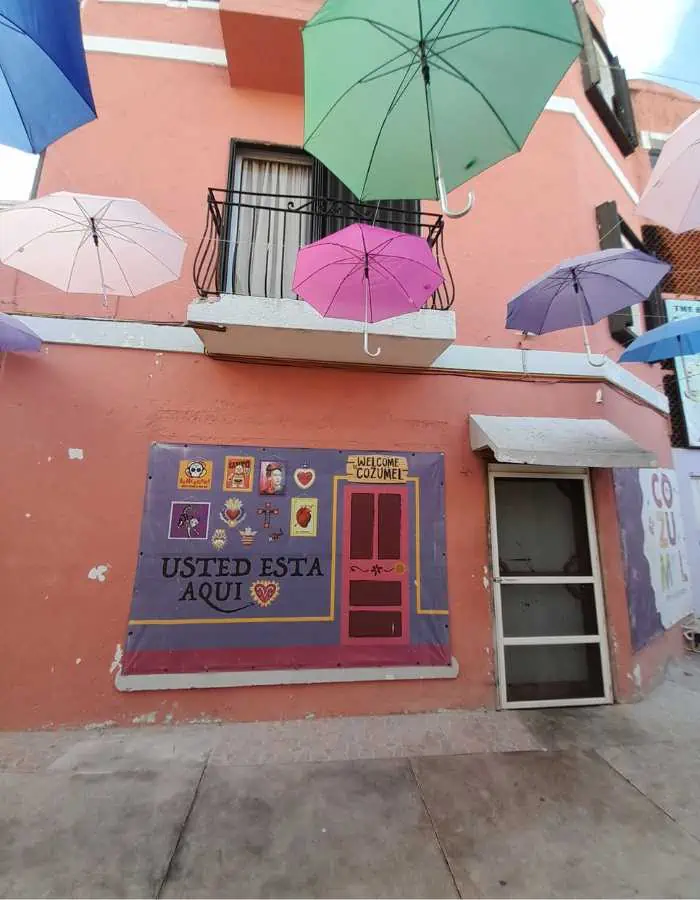 This image showcases a vibrant street scene with a pinkish-orange building adorned with a mural that reads **"USTED ESTÁ AQUÍ"** (Spanish for "You Are Here"). Above, colorful umbrellas are suspended in the air, creating a whimsical and artistic atmosphere. The mural features a stylized map or design, adding to the charm of this picturesque urban spot in Cozumel, Mexico.