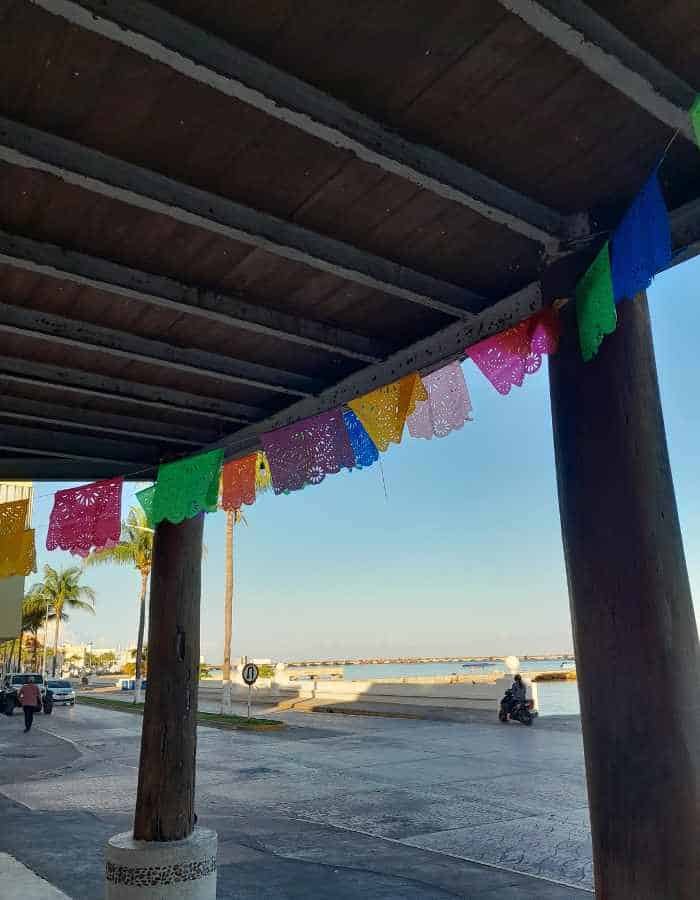 Colorful papel picado banners hang from a wooden beam roof along a seaside walkway. Palm trees, a scooter, and the ocean are visible in the background under a clear sky.
