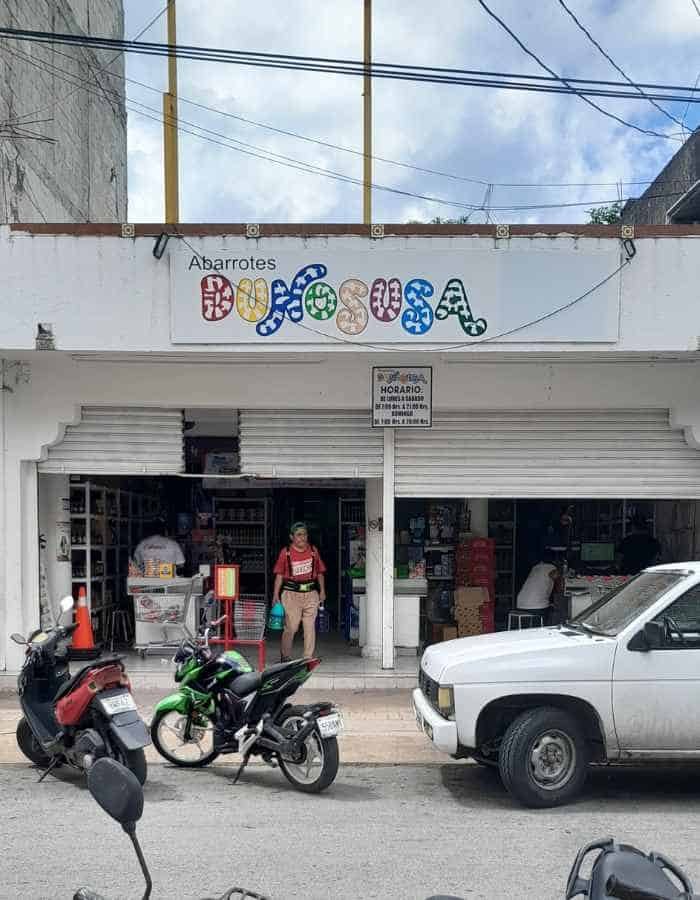 Small local grocery store named “Abarrotes Dunosusa” with an open storefront and colorful sign. Motorbikes and a white truck are parked out front, and a few people are entering or exiting under a partly cloudy sky.