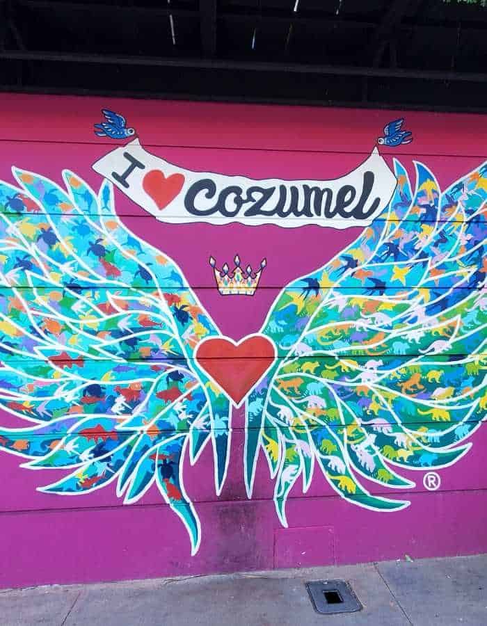 Photo of a mural featuring a pair of colorful angel wings on a bright pink wall with the phrase "I heart Cozumel" written above in a decorative banner. The wings are adorned with intricate patterns of blue, green, and yellow, framing a red heart with a golden crown at the center. Designed as a photo spot, this artwork captures the lively and artistic spirit of Cozumel, Mexico.