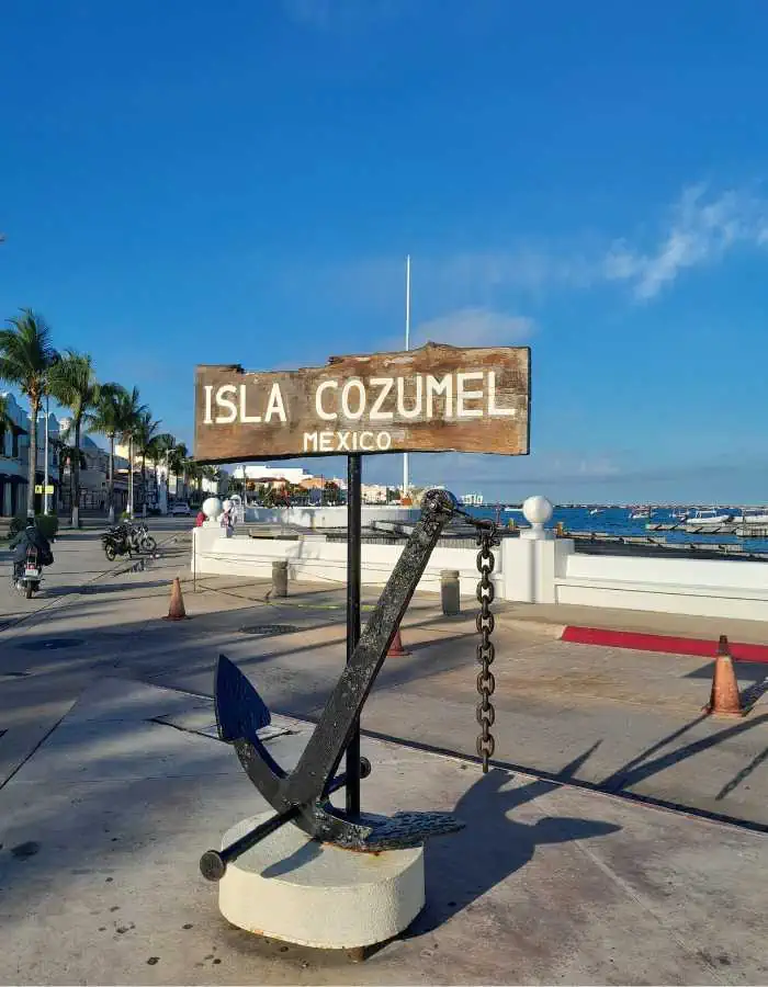 A wooden sign reading “Isla Cozumel Mexico” mounted above a large black anchor sculpture. The sign is located on a waterfront promenade with palm trees, white railings, and the ocean in the background. The scene is bright and sunny, evoking a coastal island atmosphere.