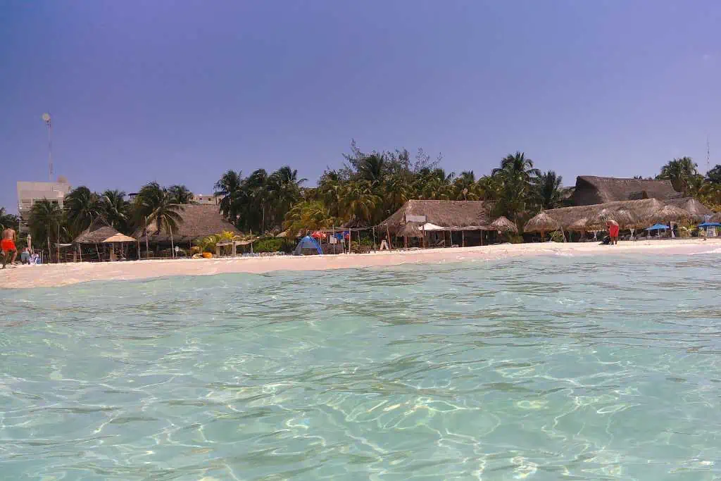A view from shallow turquoise water looking toward a sandy beach lined with palm trees and thatched-roof huts. The water is clear and calm, reflecting sunlight. The beach appears relaxed and tropical, with a few colorful umbrellas visible in the distance.