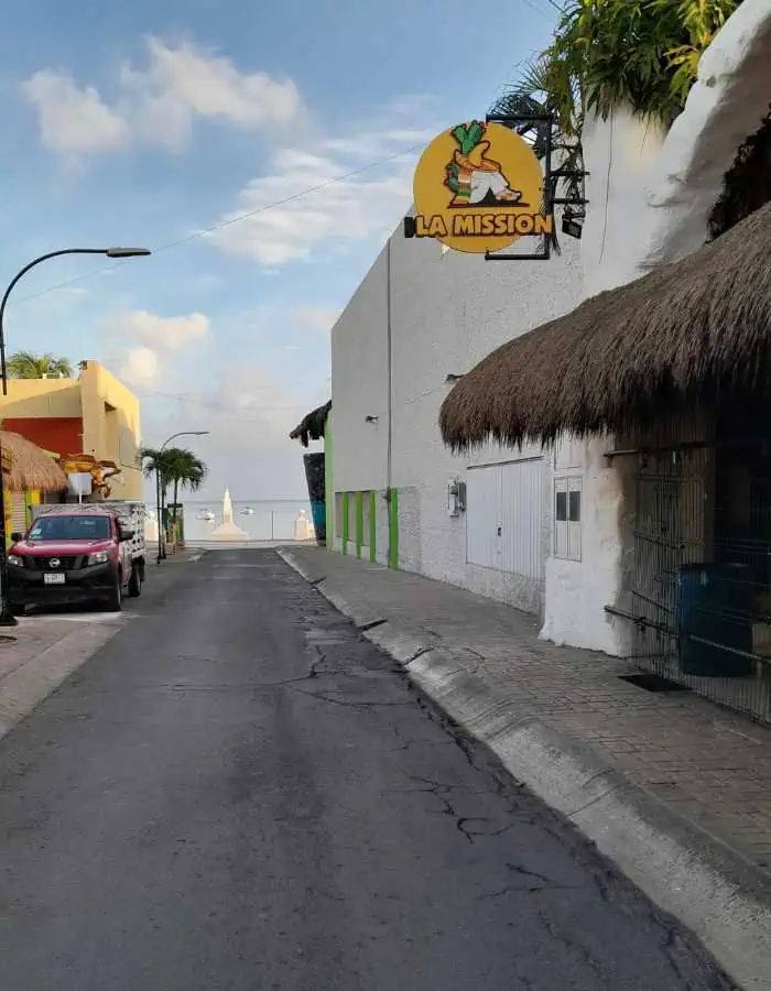 This image captures a quiet street in Cozumel, Mexico, leading toward the ocean in the background. On the right side, a thatched-roof building features a sign for "La Mission," a well-known restaurant with a pineapple logo. The scene is bathed in soft daylight, with a few parked cars and tropical greenery adding to the relaxed island atmosphere.