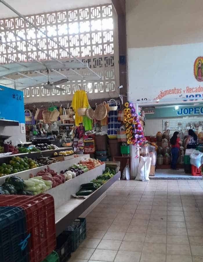 Indoor market scene with fresh produce displayed in crates and baskets, including onions, peppers, and greens. Stalls in the background sell hats, baskets, and other goods, with shoppers browsing under natural light filtering through decorative concrete walls.