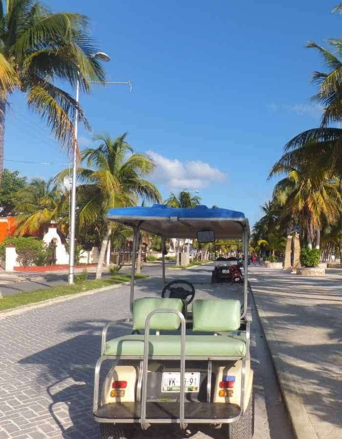 A golf cart on the street in Isla Mujeres. Palm trees line the streets and the sky is blue with a few puffy clouds.