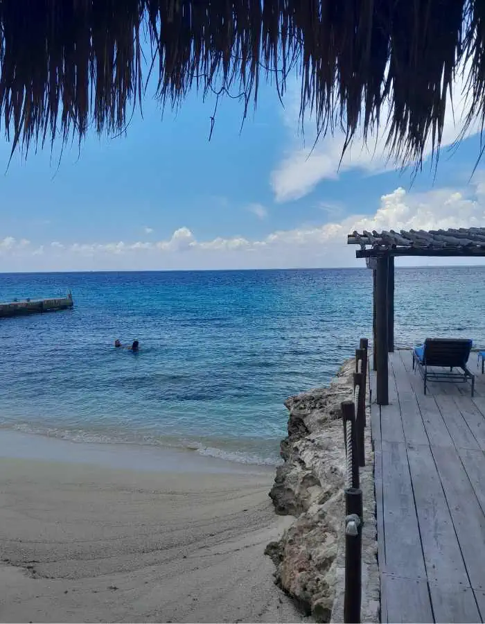 A peaceful beach scene at Playa Azul in Cozumel, with two people swimming in the calm turquoise water, a small sandy cove, and a shaded wooden deck with lounge chairs overlooking the sea. Palm thatch and a pergola frame the view under a partly cloudy sky.