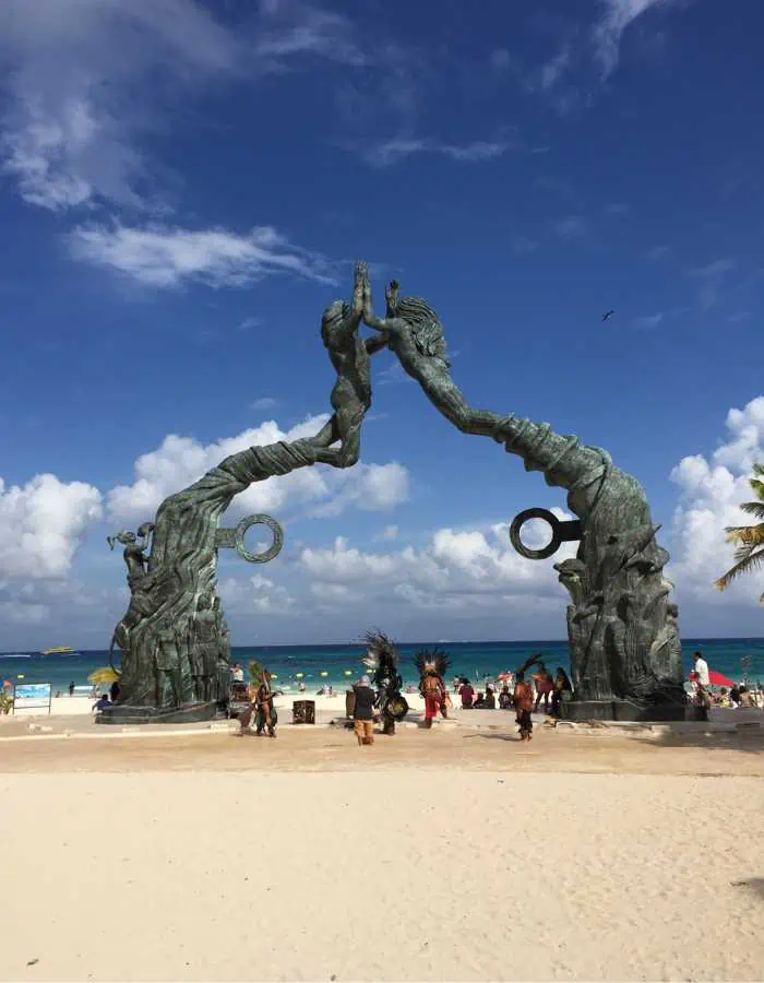 Visitors gather around the iconic 'Portal Maya' sculpture on the sandy beach of Playa del Carmen under a bright blue sky. (How to get from Tulum to Playa del Carmen)
