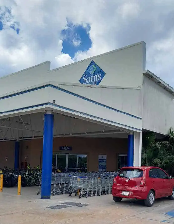 Exterior of a Sam’s Club store with shopping carts lined up near the entrance and a red car parked in front. The building features blue support columns and a large logo under a partly cloudy sky.