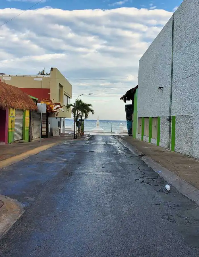 A narrow street leads toward the ocean, framed by simple buildings with colorful accents. A lone palm tree stands at the end of the road near the sandy beach and water.