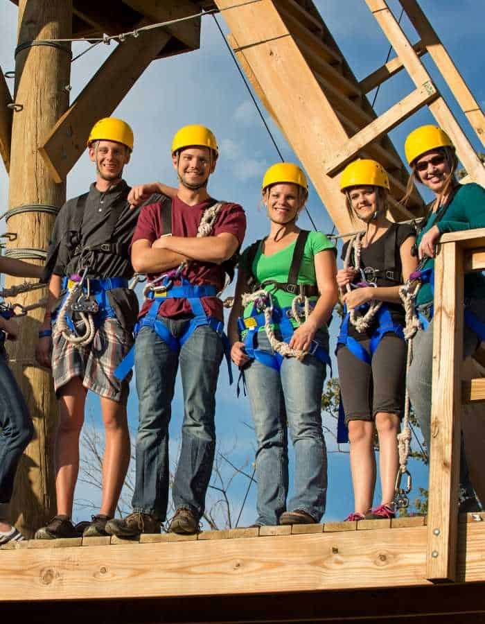 Group of five people wearing yellow helmets and harnesses stand on a wooden platform, ready for a zip-lining or ropes course adventure. They smile at the camera under a clear blue sky.