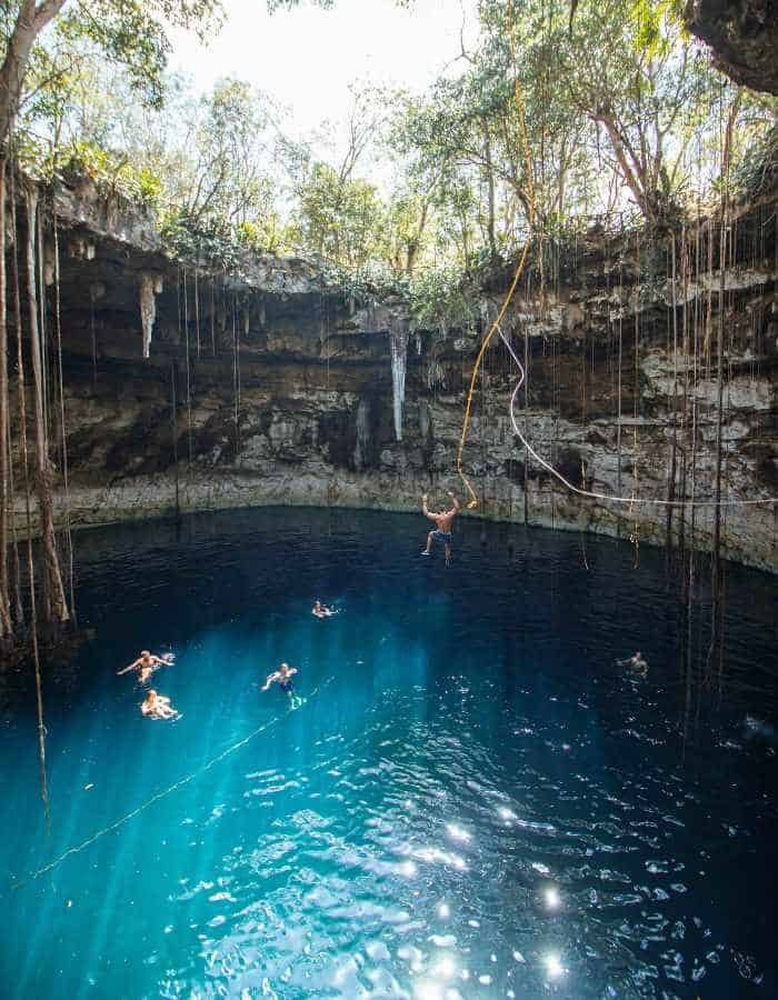 Swimmers enjoy a deep blue cenote surrounded by rocky walls and hanging vines. Sunlight filters through the opening above, casting bright rays onto the water’s surface.