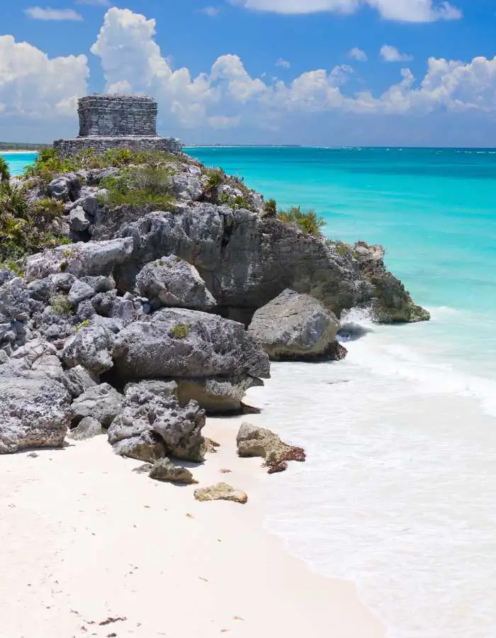 Ancient stone ruin sits atop rocky cliffs overlooking turquoise ocean water and white sand beach. Bright sunlight and scattered clouds enhance the tropical coastal scenery.