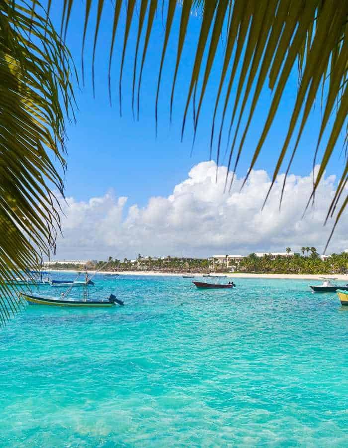 View of turquoise ocean water framed by palm fronds, with small boats floating near the shore. A distant coastline with palm trees sits under a bright blue sky with fluffy white clouds.