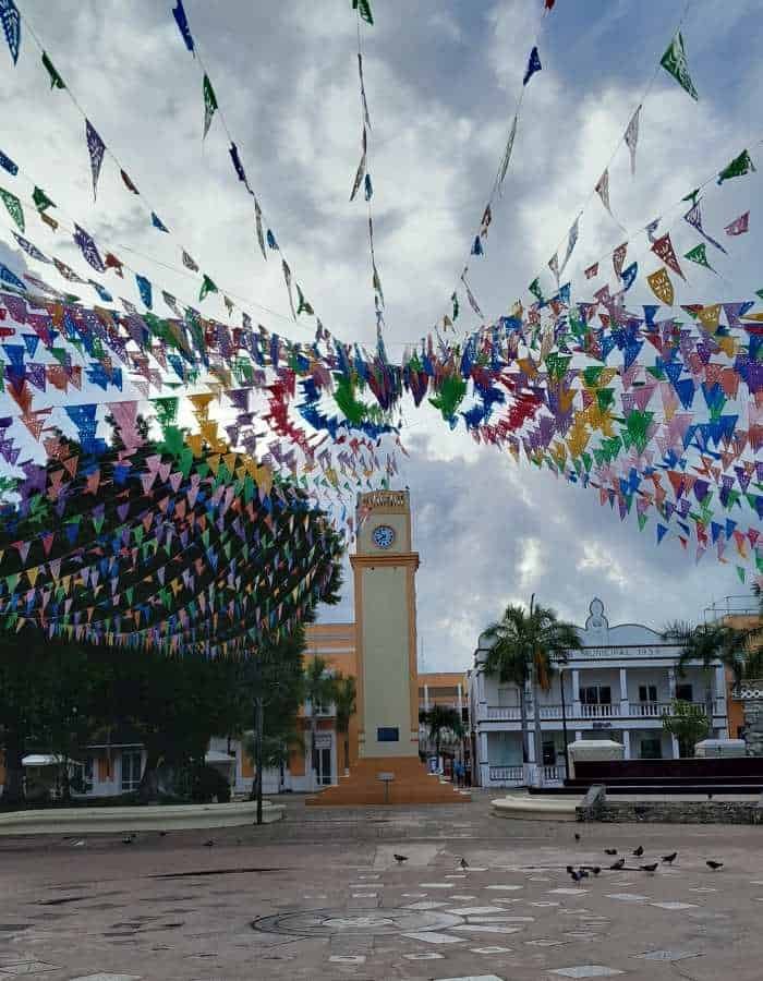A town square decorated with vibrant strings of multicolored triangular flags radiating from a central clock tower. The scene captures a festive atmosphere under a cloudy sky, with colonial-style buildings and palm trees in the background and a few birds on the ground.