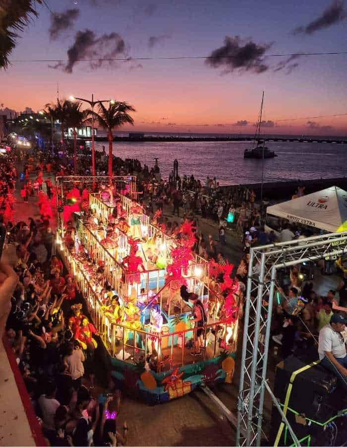 Vibrant street parade along a seaside boulevard at sunset, featuring a brightly lit float decorated with colorful lights and performers in red costumes. Crowds of people line the street, enjoying the festive atmosphere against a backdrop of palm trees, ocean, and a glowing sky.