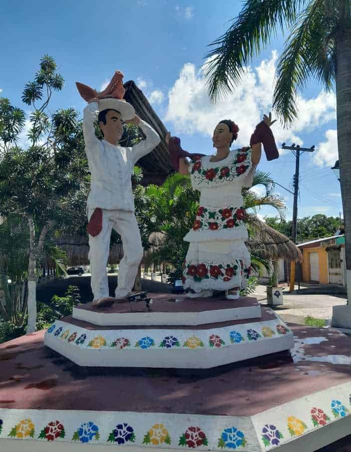 Colorful statue of a traditional Mexican couple dancing, with the man in a white outfit and sombrero and the woman in a white dress adorned with bright floral embroidery. The sculpture stands on a decorated pedestal surrounded by palm trees, evoking cultural pride and festive spirit in a tropical village setting.