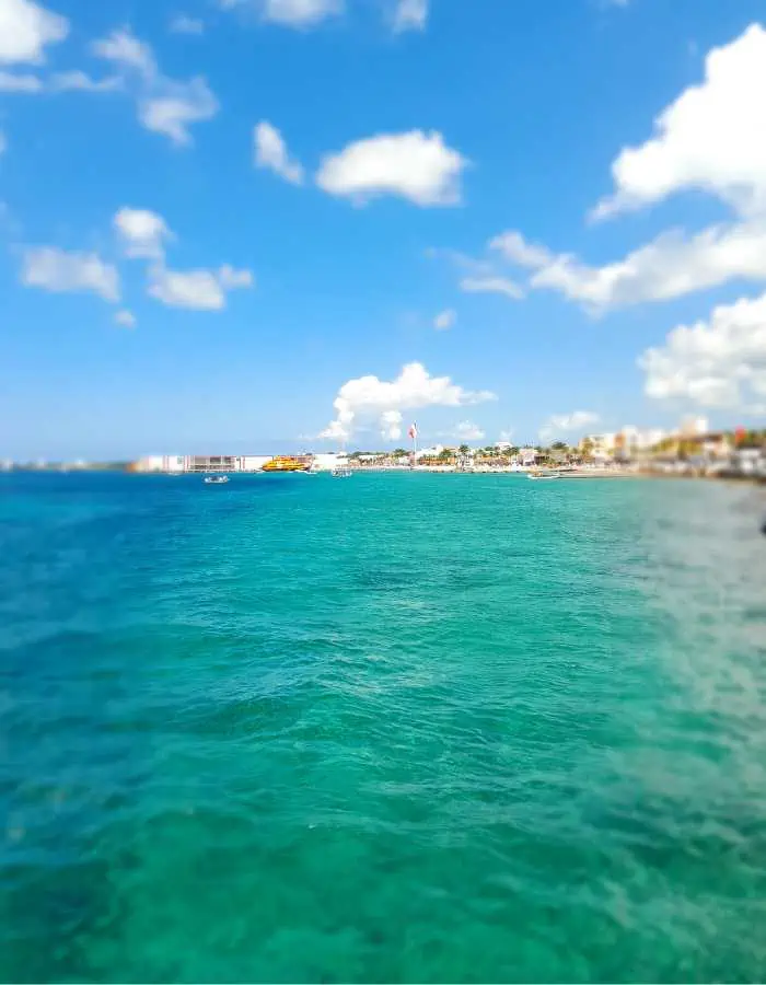 Bright and sunny coastal view showcasing turquoise-blue ocean water stretching toward a distant shoreline with buildings and palm trees. The image captures the vibrant colors and clarity of a tropical beach destination under a sky dotted with fluffy white clouds.