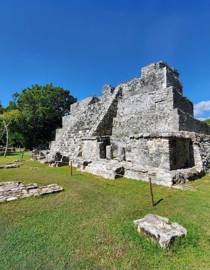 Ancient stone pyramid-style ruins at El Rey Archaeological Zone in Cancún, surrounded by grassy terrain and trees, with a clear blue sky overhead.