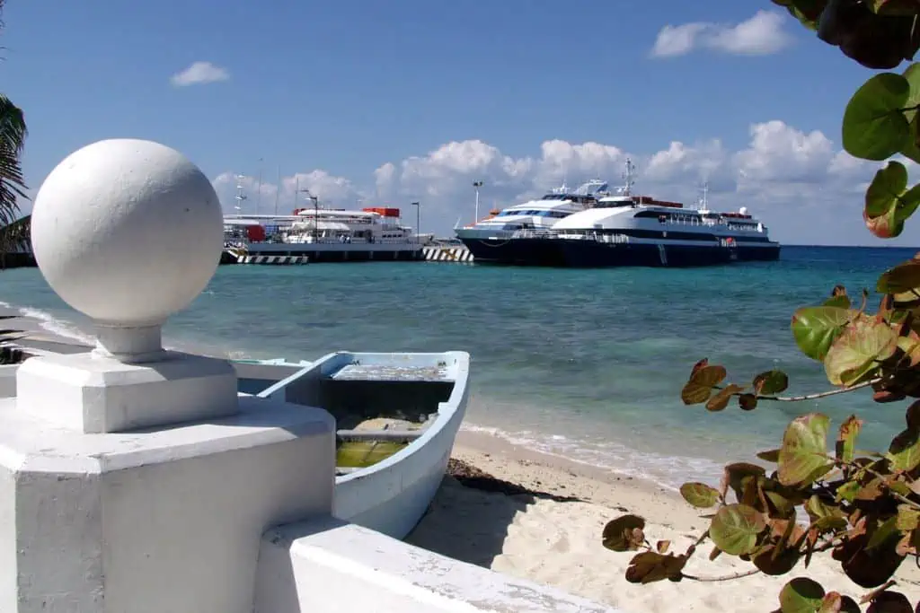 View from a sandy shore shows a small boat near a white concrete railing in the foreground, with large ferries docked at a pier in the turquoise water. Bright blue sky and scattered clouds complete the peaceful coastal scene.