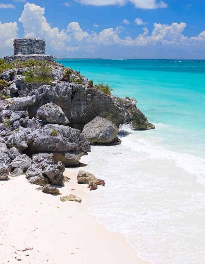 Ancient stone ruin sits atop rocky cliffs overlooking turquoise waters and a pristine white sand beach. Bright sunlight and scattered clouds enhance the tropical coastal scenery.