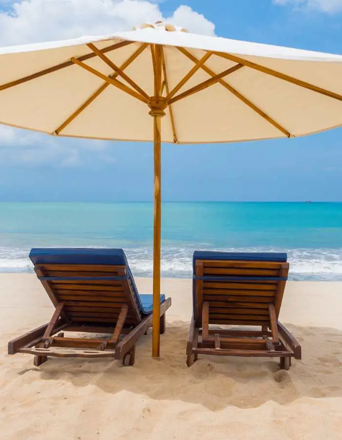 Two wooden lounge chairs with navy blue cushions sit under a large white beach umbrella facing the turquoise ocean on a sandy beach under a sunny sky.