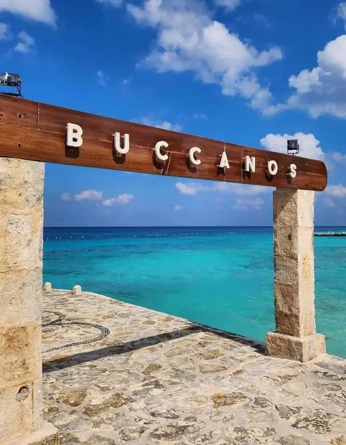 A wooden and stone entrance sign for "Buccanos" stands against a stunning turquoise ocean backdrop in Cozumel, Mexico. The clear blue sky with fluffy white clouds enhances the tropical beach setting. The stone walkway beneath the sign leads toward the water, inviting visitors to this picturesque seaside destination.