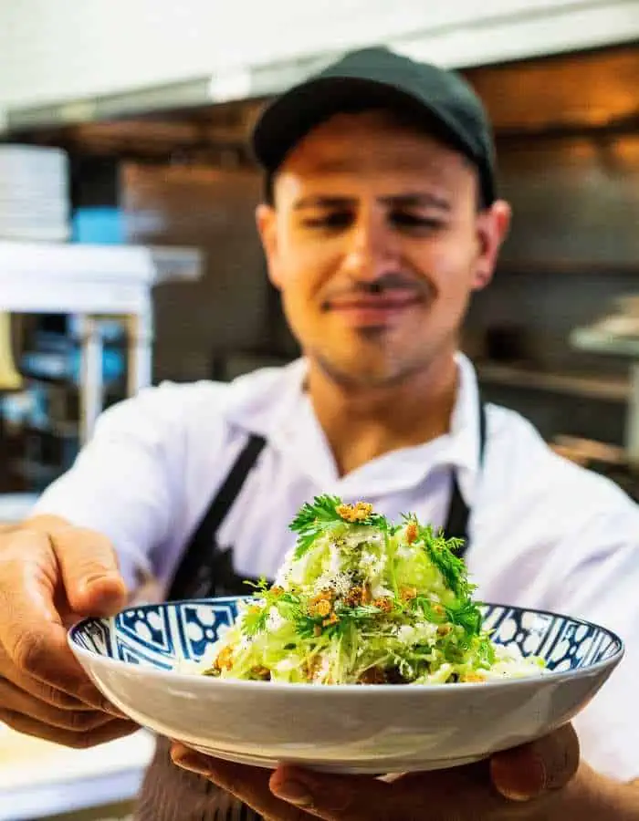 Smiling waiter in a black cap and white shirt presenting a bowl of fresh guacamole topped with herbs and crumbled cheese in a decorative blue-and-white bowl, inside a restaurant kitchen.