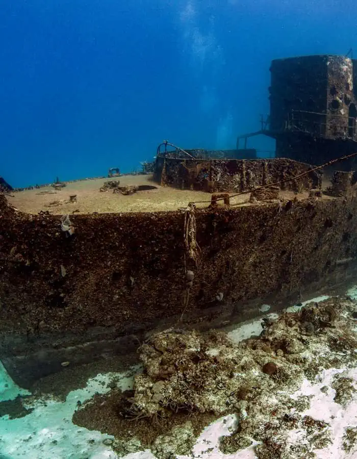 A sunken shipwreck rests on the ocean floor in Cozumel, Mexico, covered in coral growth and marine life. The rusted hull and towering structure of the ship create an eerie yet fascinating underwater scene. The clear blue water provides excellent visibility, making this wreck a captivating sight for divers and submarine passengers.