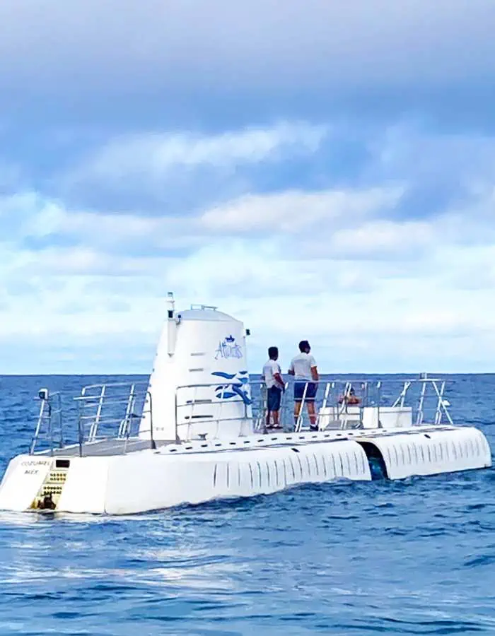 The Atlantis Submarine floats on the surface of the ocean in Cozumel, Mexico, with crew members standing on top, preparing for a dive. The sleek white vessel contrasts against the deep blue sea and a partly cloudy sky. Gentle waves ripple around the submarine, setting the scene for an exciting underwater adventure.