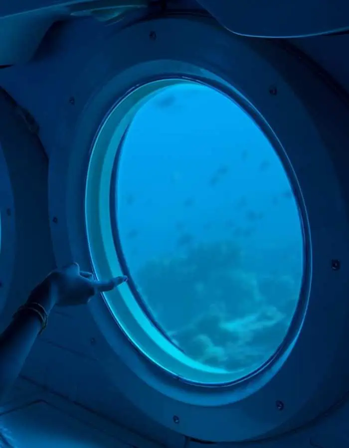A passenger's hand reaches toward the circular window of the Atlantis Submarine in Cozumel, Mexico, as they gaze at the underwater world outside. The deep blue ocean is filled with fish swimming near the coral reef, creating a mesmerizing view.