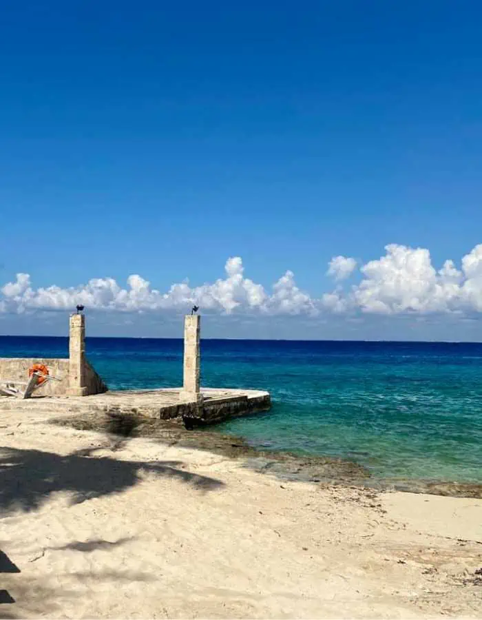 A peaceful beach scene at Buccanos in Cozumel, Mexico, featuring a sandy shoreline with a small stone pier extending into the clear turquoise waters. The deep blue ocean meets the horizon under a bright blue sky with fluffy white clouds.