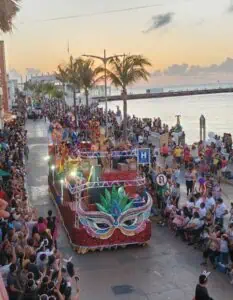 A vibrant parade float decorated with a large, colorful carnival mask moves along a waterfront street lined with palm trees and packed with cheering spectators. The festive scene unfolds at sunset, with dancers in costume and lights adding to the lively coastal celebration.