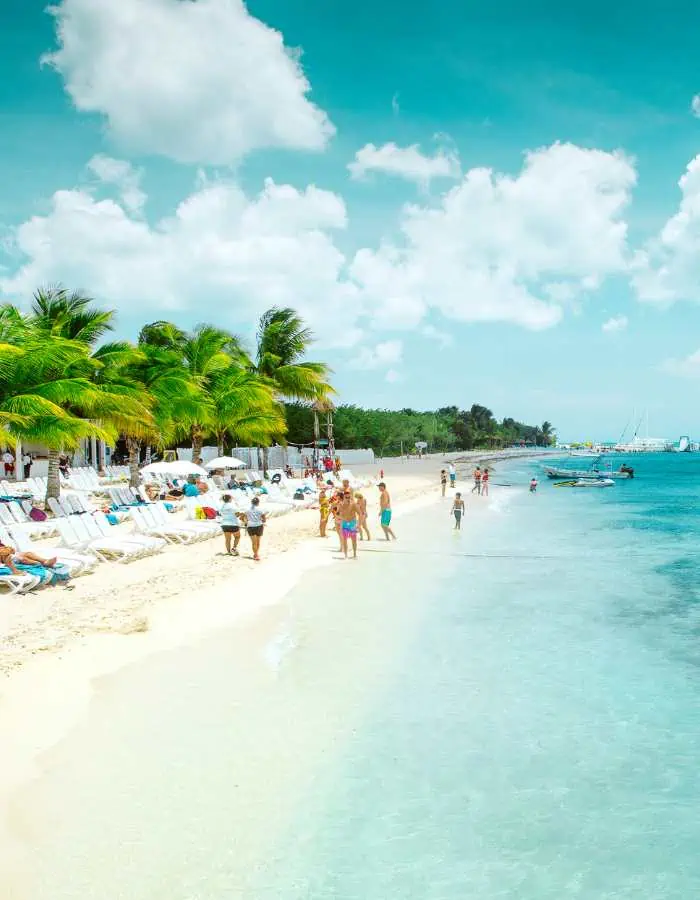 Tourists stroll along a stunning beach in Cozumel lined with palm trees, white lounge chairs, and clear turquoise water under a bright sky with scattered clouds.