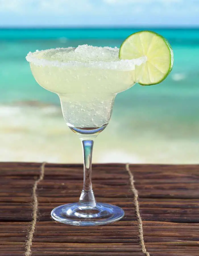 Classic frozen margarita in a salt-rimmed glass garnished with a lime wheel, set on a wooden table with the turquoise ocean and sandy beach blurred in the background.