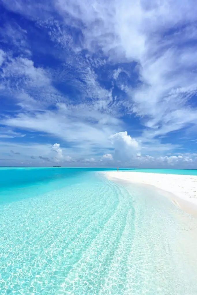 Crystal-clear turquoise water gently laps against a pristine white sandbar under a vibrant blue sky filled with wispy clouds. The tranquil tropical scene stretches to the horizon, creating a sense of peaceful isolation.