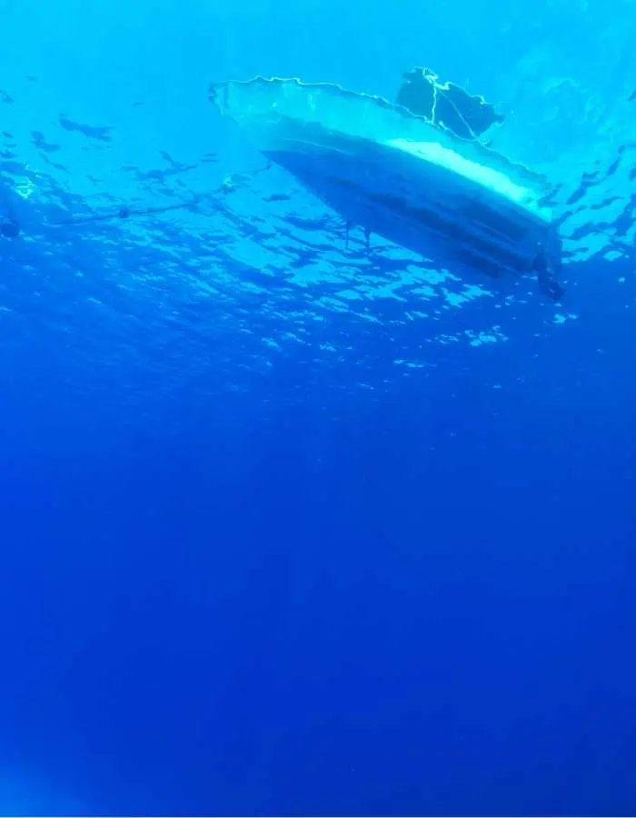 A small boat is seen from underwater, floating at the surface of a deep blue ocean. Sunlight filters through the water, creating rippling light patterns above.