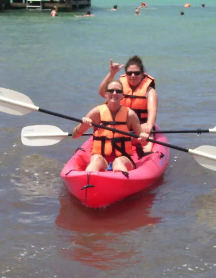 Two women wearing orange life vests kayaking in a red tandem kayak on the shallow shoreline of Paradise Beach, with clear turquoise water and other beachgoers in the background.