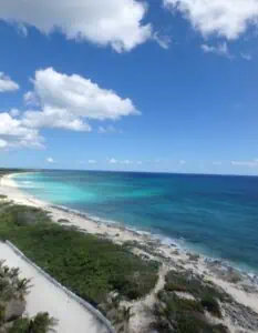 A sweeping aerial view of a remote coastline shows white sandy beach, turquoise and deep blue ocean, and patches of green vegetation under a bright blue sky with scattered clouds. The natural landscape appears untouched and serene.