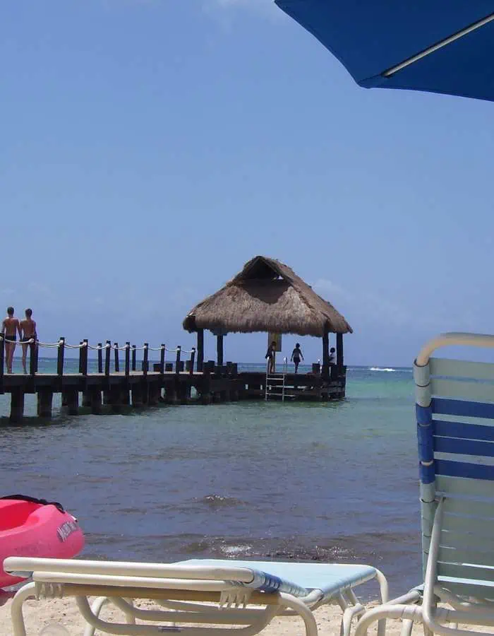 Relaxing beach scene with lounge chairs in the foreground, a thatched-roof pier extending into turquoise waters, and a few people enjoying the view under a clear blue sky.