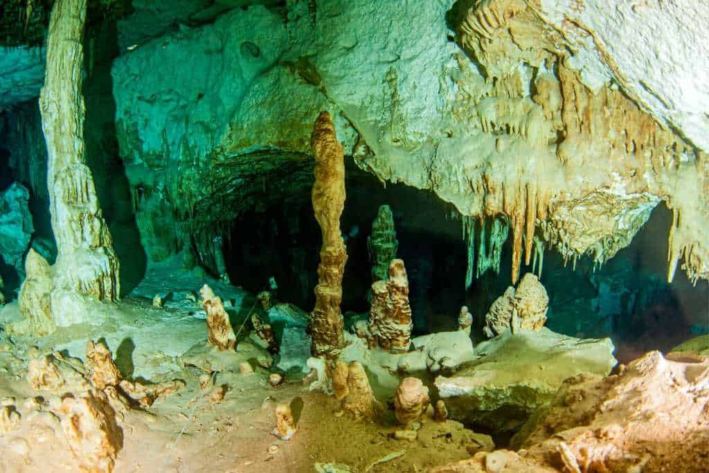 Limestone cave interior with dramatic stalactites hanging from the ceiling and stalagmites rising from the floor. The cave is dimly lit with green and amber tones, highlighting its natural formations and rugged textures.