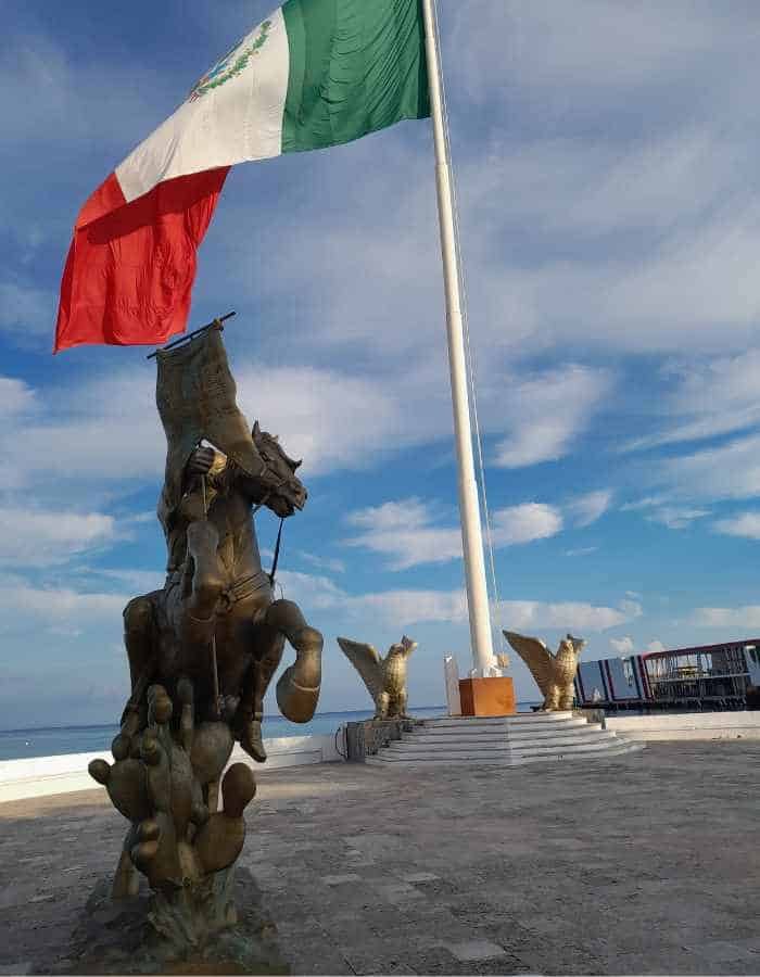 Bronze statue of an eagle on a cactus with a snake in its beak stands prominently in front of a massive Mexican flag waving on a tall pole. The monument overlooks the ocean under a partly cloudy sky.