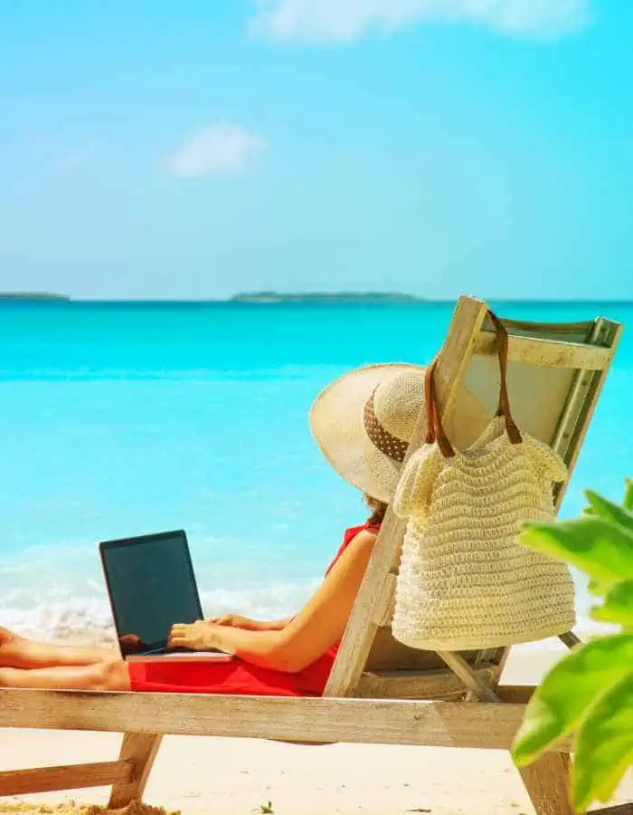 A woman in a sunhat and red dress lounges on a beach chair with a laptop on her lap, facing crystal-clear turquoise water. A woven beach bag hangs from the chair, and tropical leaves frame the sunny, serene remote work scene.