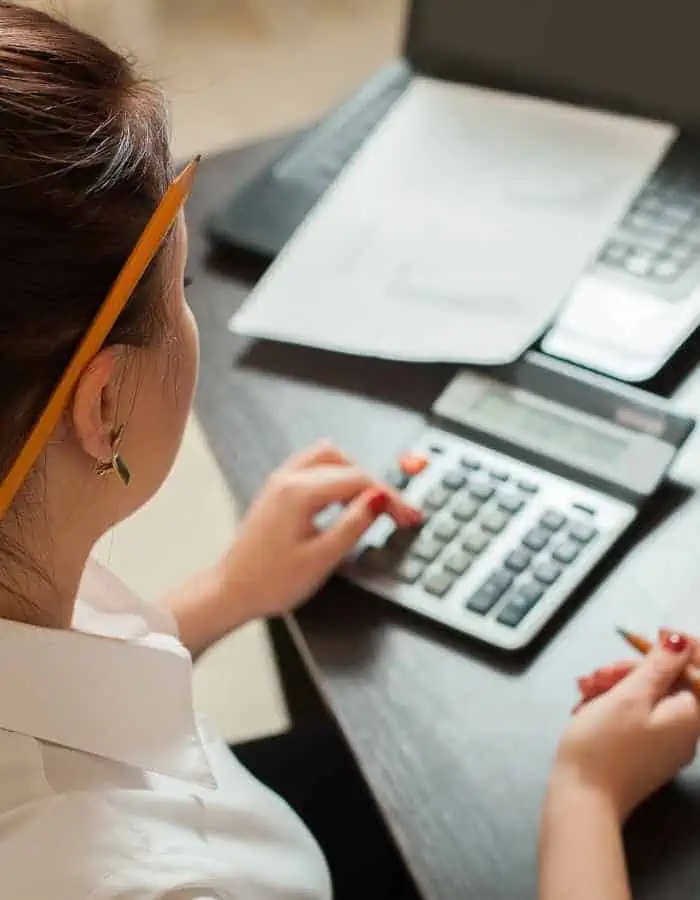 A woman with a pencil in her hair uses a calculator while working at a desk with papers and a laptop. The over-the-shoulder view suggests she’s focused on bookkeeping, accounting, or financial tasks.