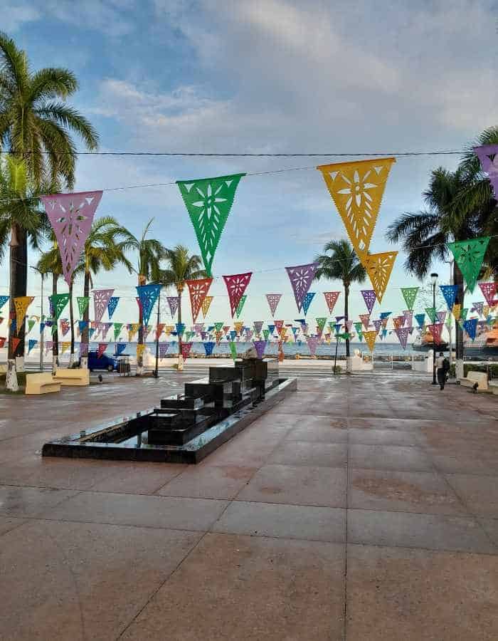 Cozumel’s main square decorated with vibrant papel picado flags in green, yellow, purple, and pink, strung above a paved plaza lined with palm trees and a central fountain. The sky is partly cloudy, and the ocean is visible in the background.