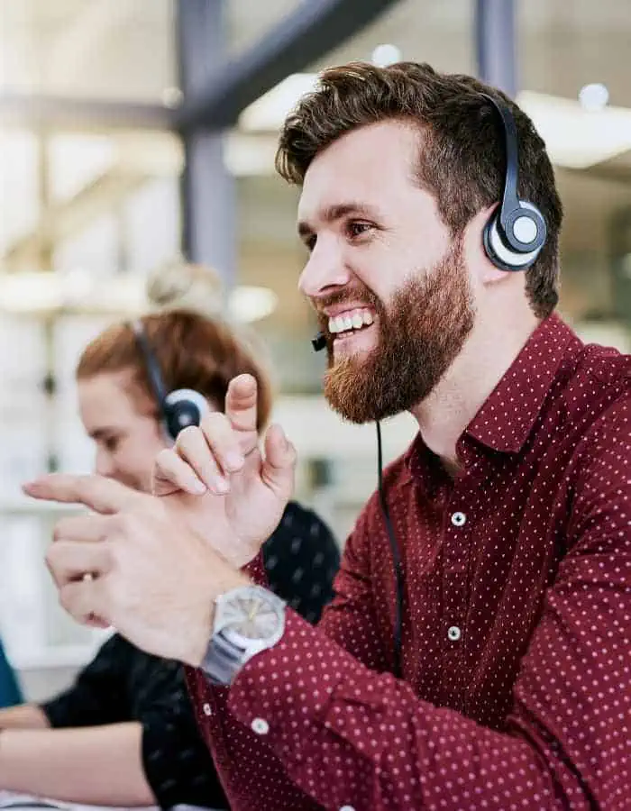 A cheerful man with a beard and headset gestures while speaking, seated in a bright office environment. He’s wearing a maroon polka-dot shirt, suggesting he’s part of a customer service or support team, with a colleague working in the background.