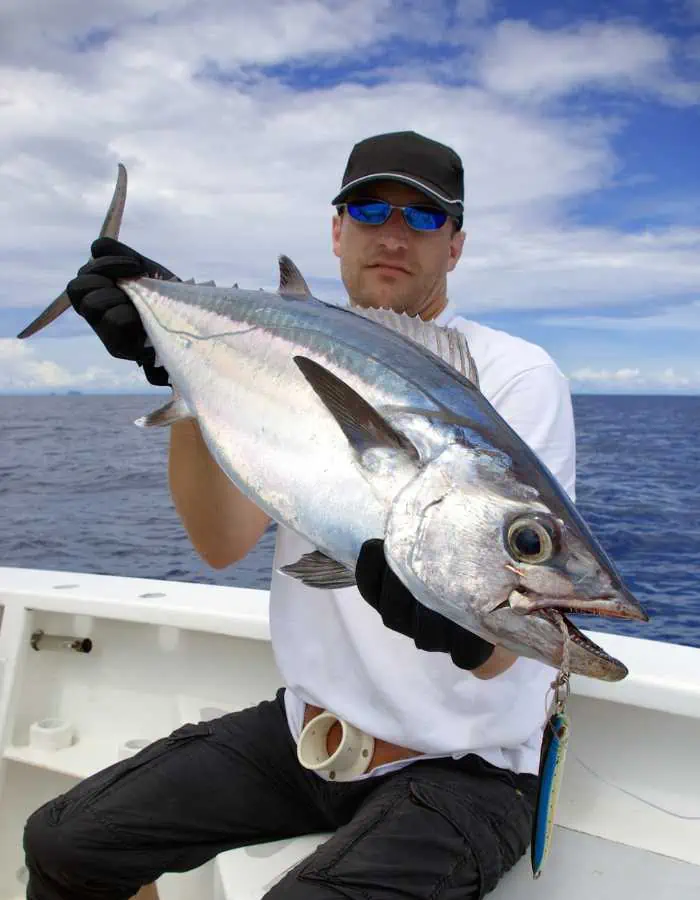 Man wearing sunglasses and gloves proudly holding a large tuna caught during a deep sea fishing trip, with the open ocean and partly cloudy sky in the background.