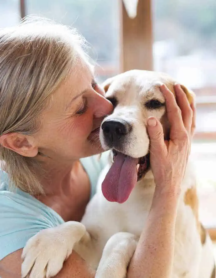 A joyful woman with blonde hair lovingly kisses a happy, panting dog on the face while gently holding its head. The close-up captures a warm, affectionate moment, perfect for illustrating pet sitting or the bond between pets and their caregivers.