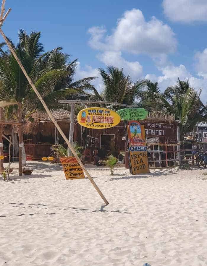 A rustic beachfront restaurant called El Pescador in Cozumel, with signs advertising “Rent Stuff,” “Drinks to a Tour,” and souvenirs, set against a backdrop of palm trees and a bright blue sky. The sandy beach stretches out in front of the open-air wooden building.