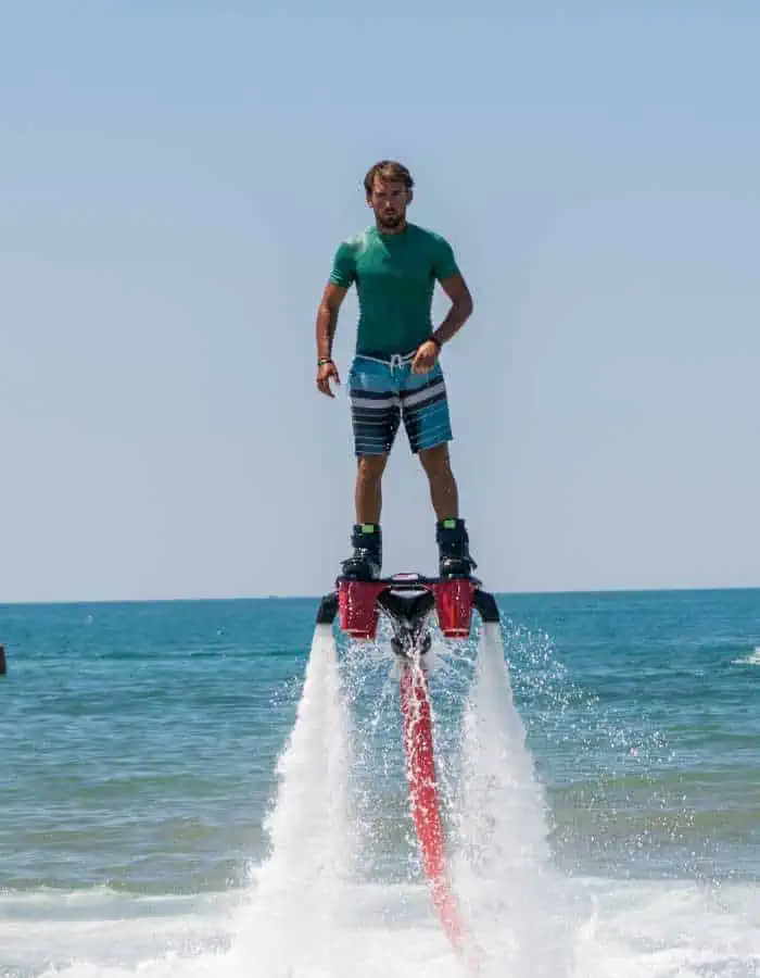 Man flyboarding above the ocean, propelled by powerful water jets from a board attached to his feet, wearing a green shirt and striped swim trunks against a clear blue sky.